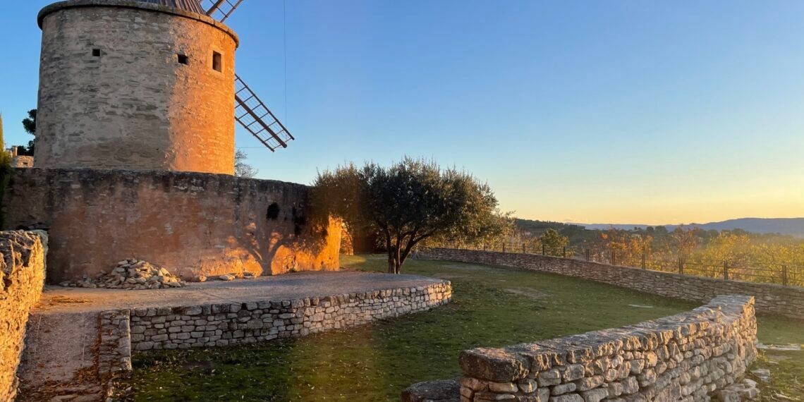 Stone windmill with sunset lighting on a rustic landscape, surrounded by stone walls and trees.